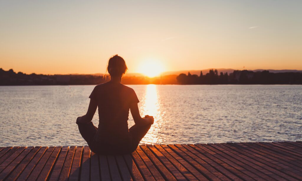 woman doing meditation and breathing exercises on wooden pier near lake, silhouette at sunrise