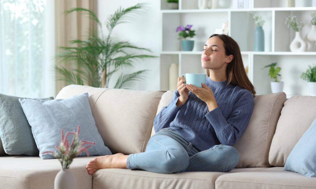 Relaxed woman in blue smelling coffee aroma sitting on a couch at home meditating for 5 minutes