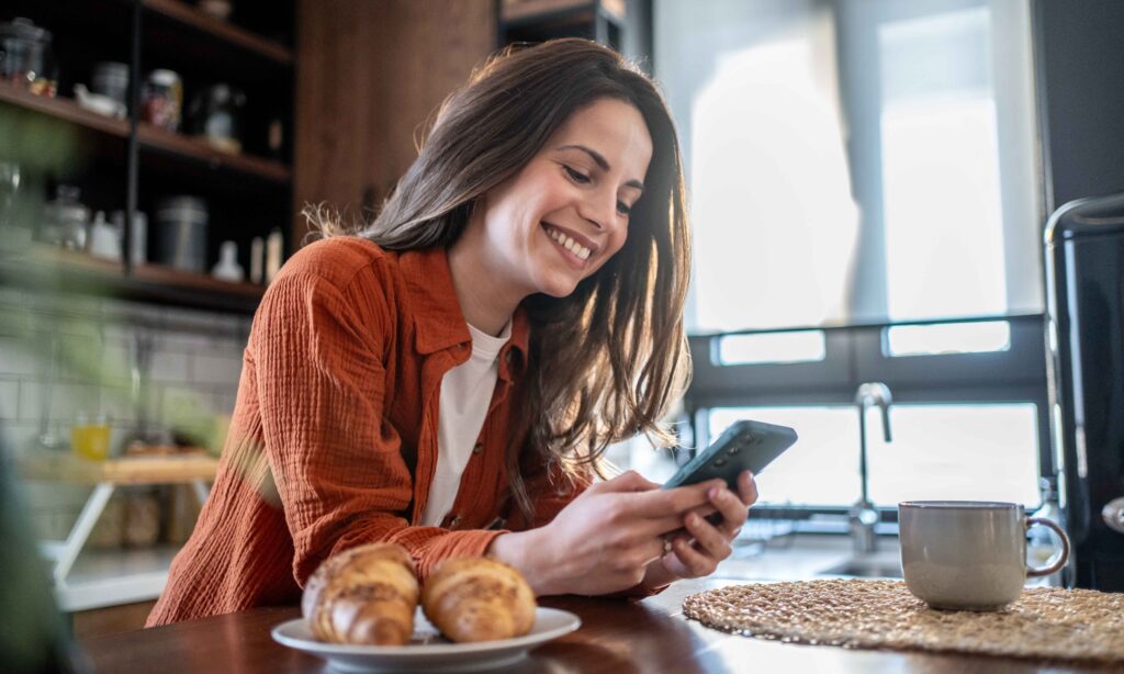 Cheerful young woman enjoying breakfast while using her smartphone, smiling brightly in a modern kitchen filled with croissants and coffee, creating a cozy morning atmosphere
