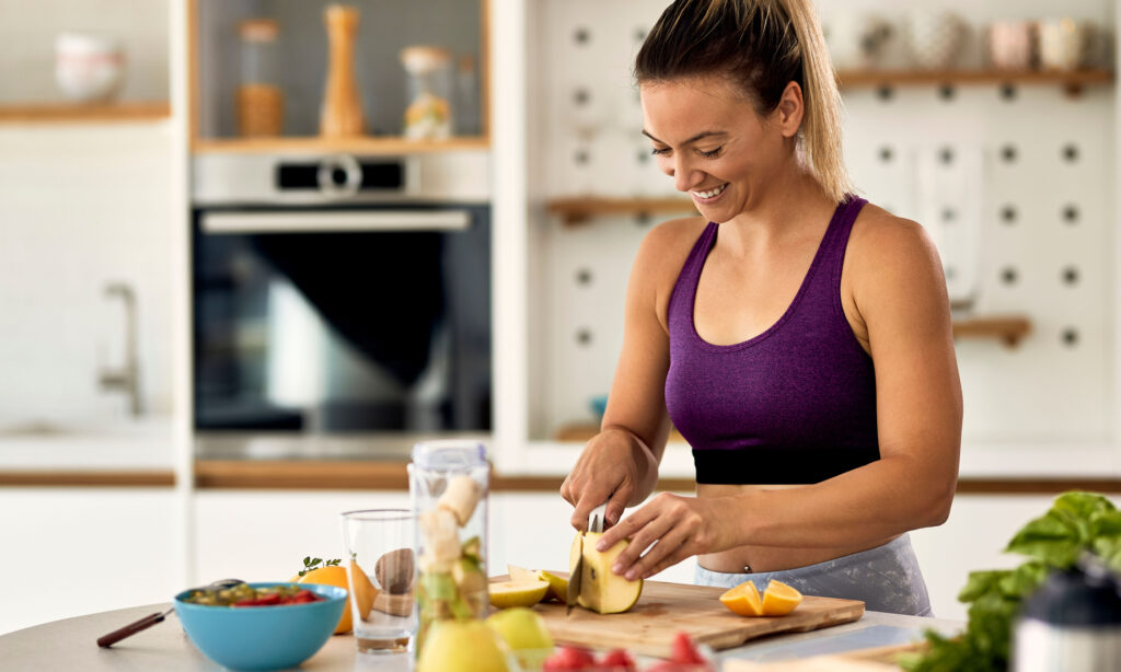 Young happy sportswoman slicing fruit while making smoothie in her kitchen.